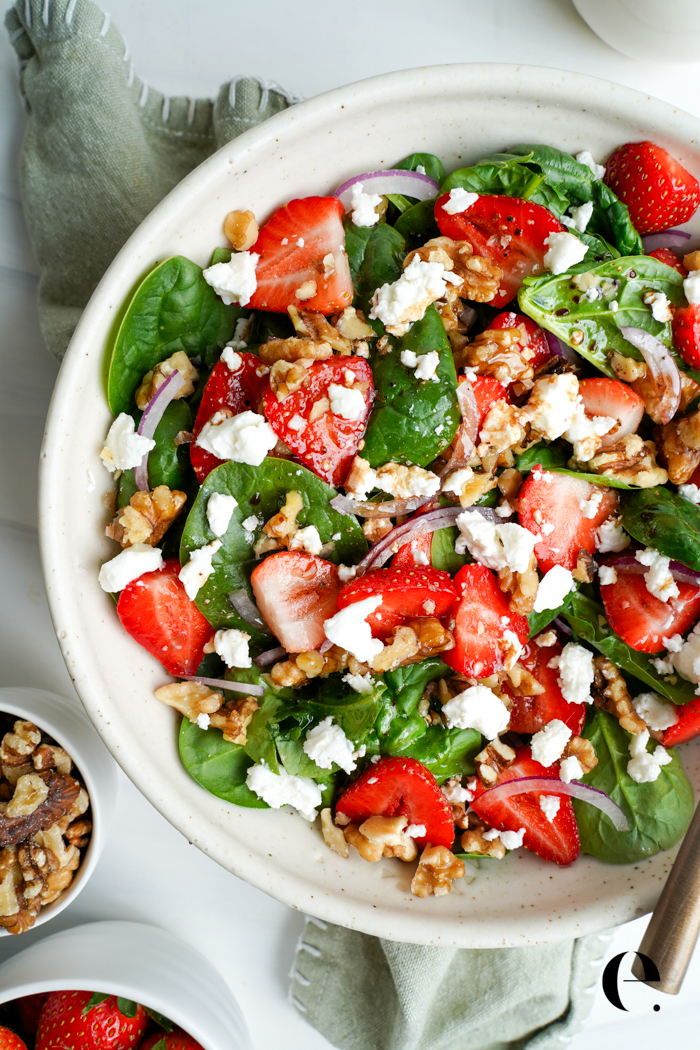 Overhead view of strawberry spinach salad with goat cheese and walnuts, with small bowls of walnuts and strawberries on the side