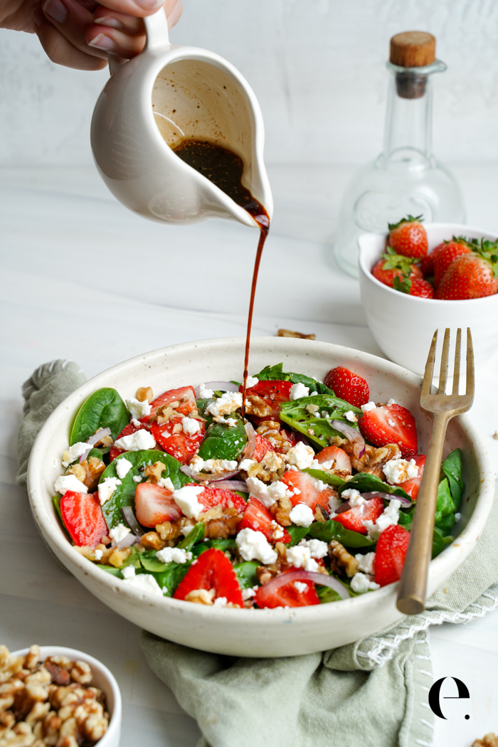 Pouring balsamic dressing over a strawberry spinach salad with goat cheese and walnuts