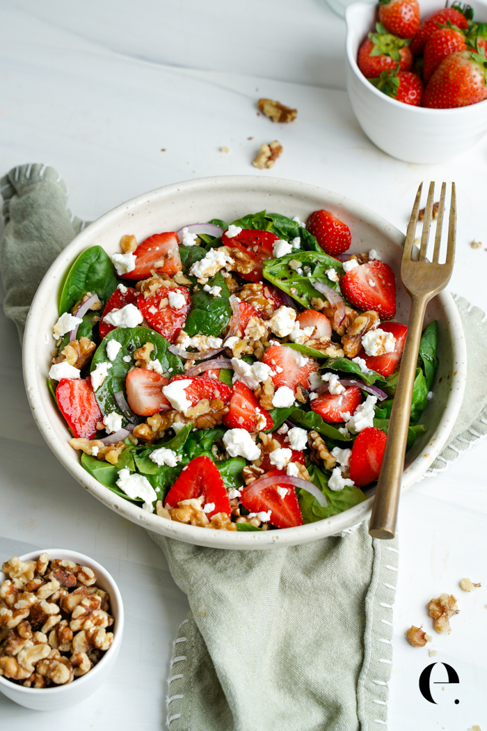 Strawberry spinach salad in a white bowl with a gold fork, with bowls of strawberries and walnuts nearby