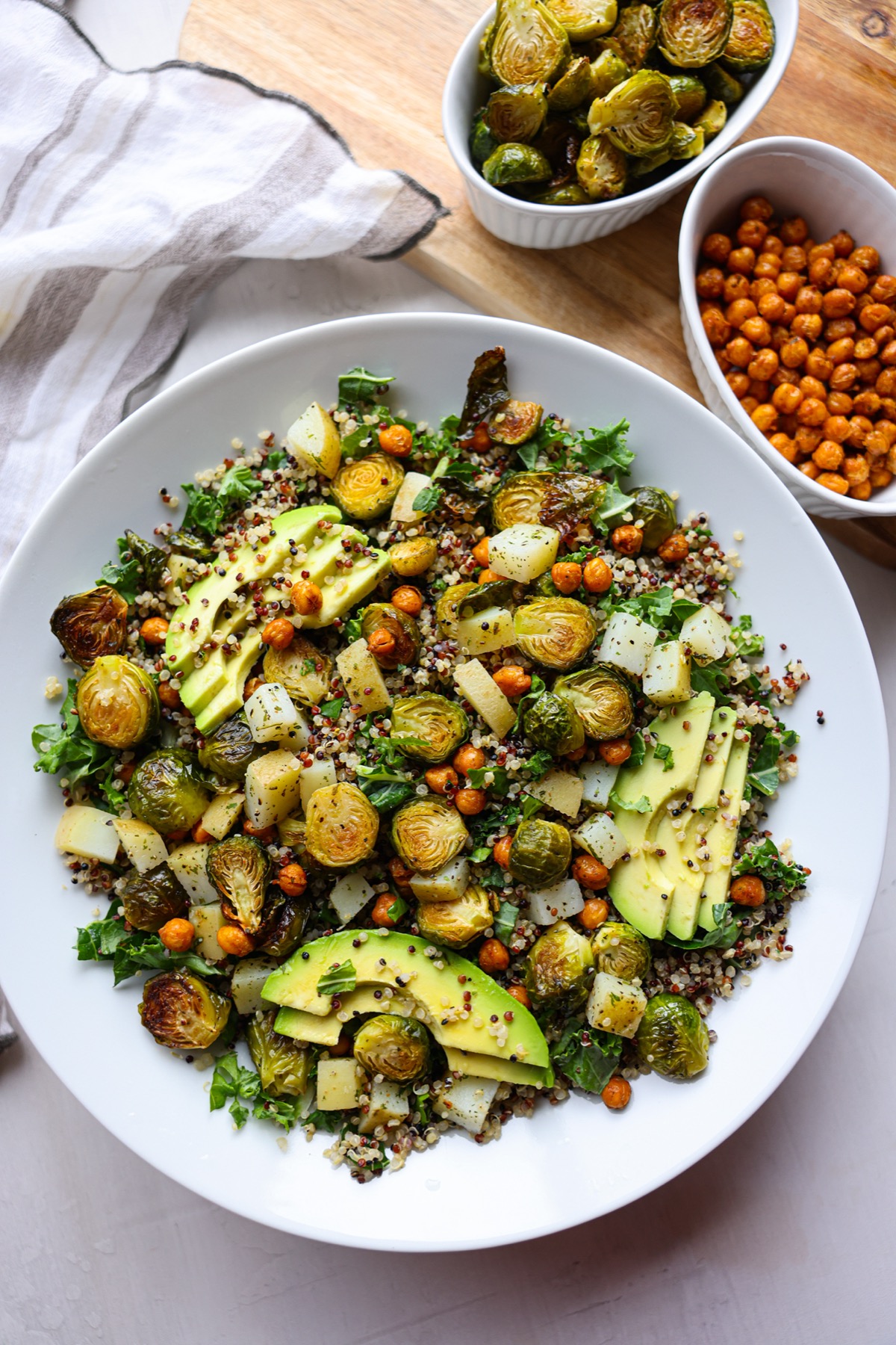 Overhead view of a Brussels sprouts quinoa bowl with chickpeas, potatoes, kale and avocado slices, next to bowls of roasted Brussels sprouts and chickpeas