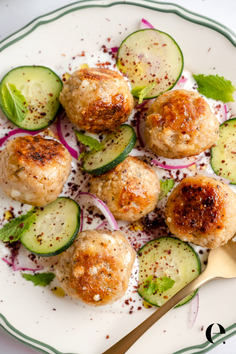 Greek chicken meatballs seared golden brown on garlic yogurt sauce with sliced cucumber, red onion, fresh mint, and sumac