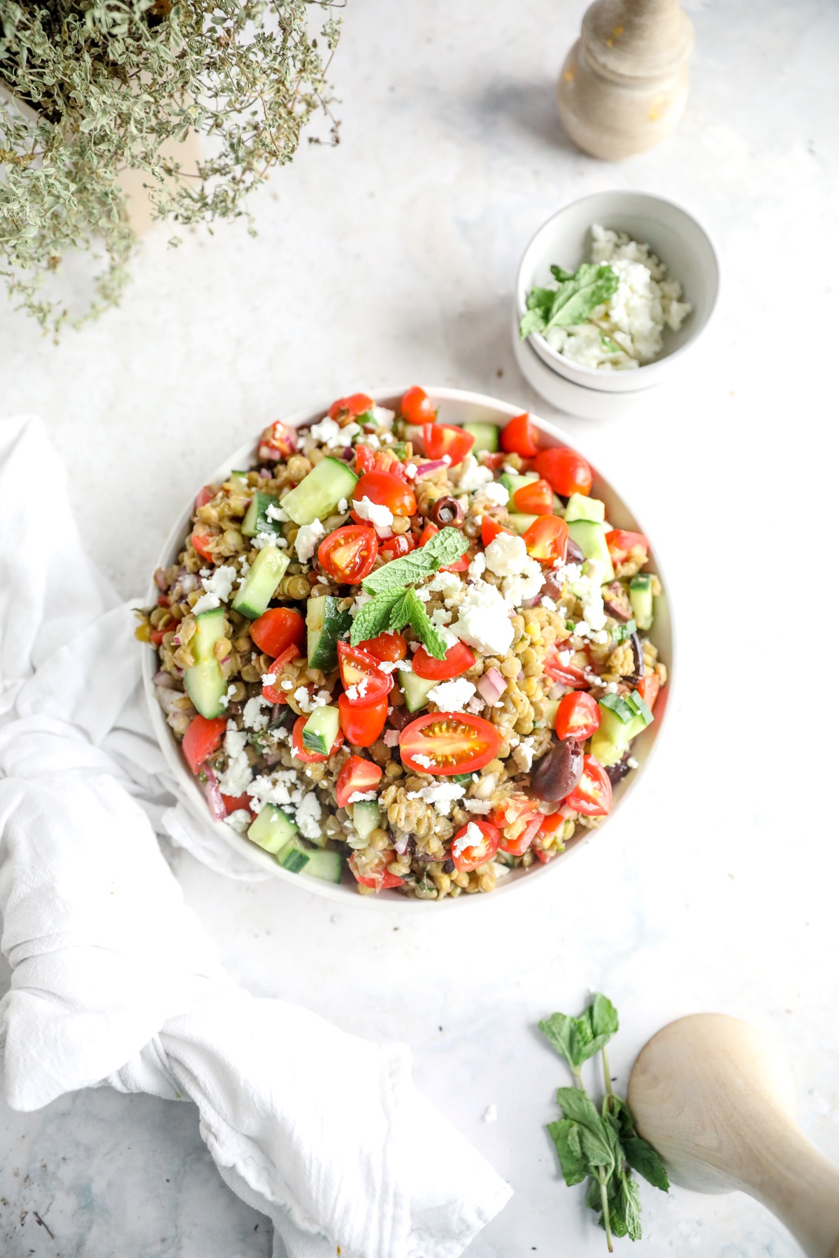 Overhead view of a bowl of green lentil salad with tomatoes, cucumber, olives and feta, next to a small bowl of feta and fresh mint