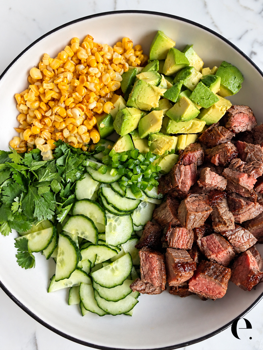 Overhead view of a steak salad with avocado and corn, with the steak, cucumber, cilantro and jalapeño arranged in sections before tossing