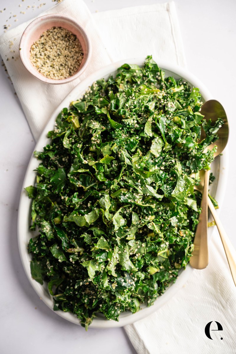 Vegan kale caesar salad on an oval platter with gold servers and a small bowl of hemp seeds